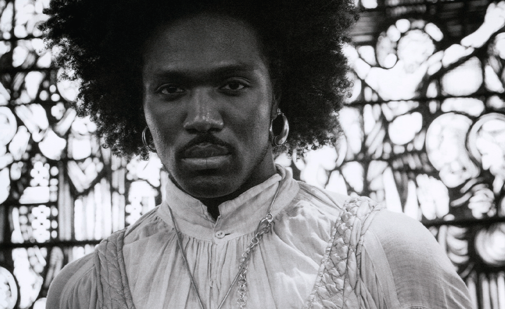 Black-and-white close-up portrait of a Black man with an afro hairstyle, moustache, and hoop earrings, looking directly into the camera. He wears a light pleated mandarin-collar shirt and necklaces, set against an intricate stained-glass window background. - Geoffrey Baptiste
