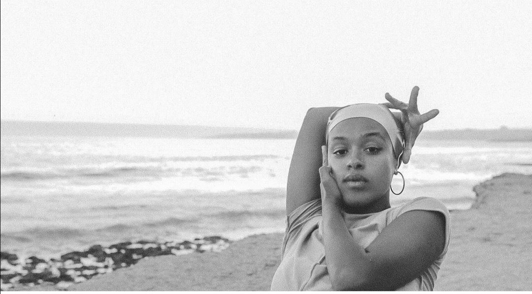 Medium black-and-white portrait of a young woman on a beach with the sea in the background. She wears a headband and hoop earrings, posing with one arm raised and her hand framing her face as she looks serenely into the camera. - Virginia Perez
