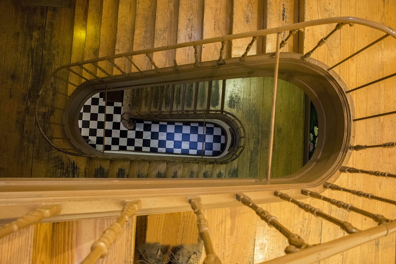 ALKANTARA - Vista em picado (de cima para baixo) através do vão central de uma escadaria espiral em madeira com um corrimão de balaústres em madeira clara. No piso inferior, no centro da perspetiva, destaca-se o chão em azulejo ou mosaico com um padrão xadrez a preto e branco.