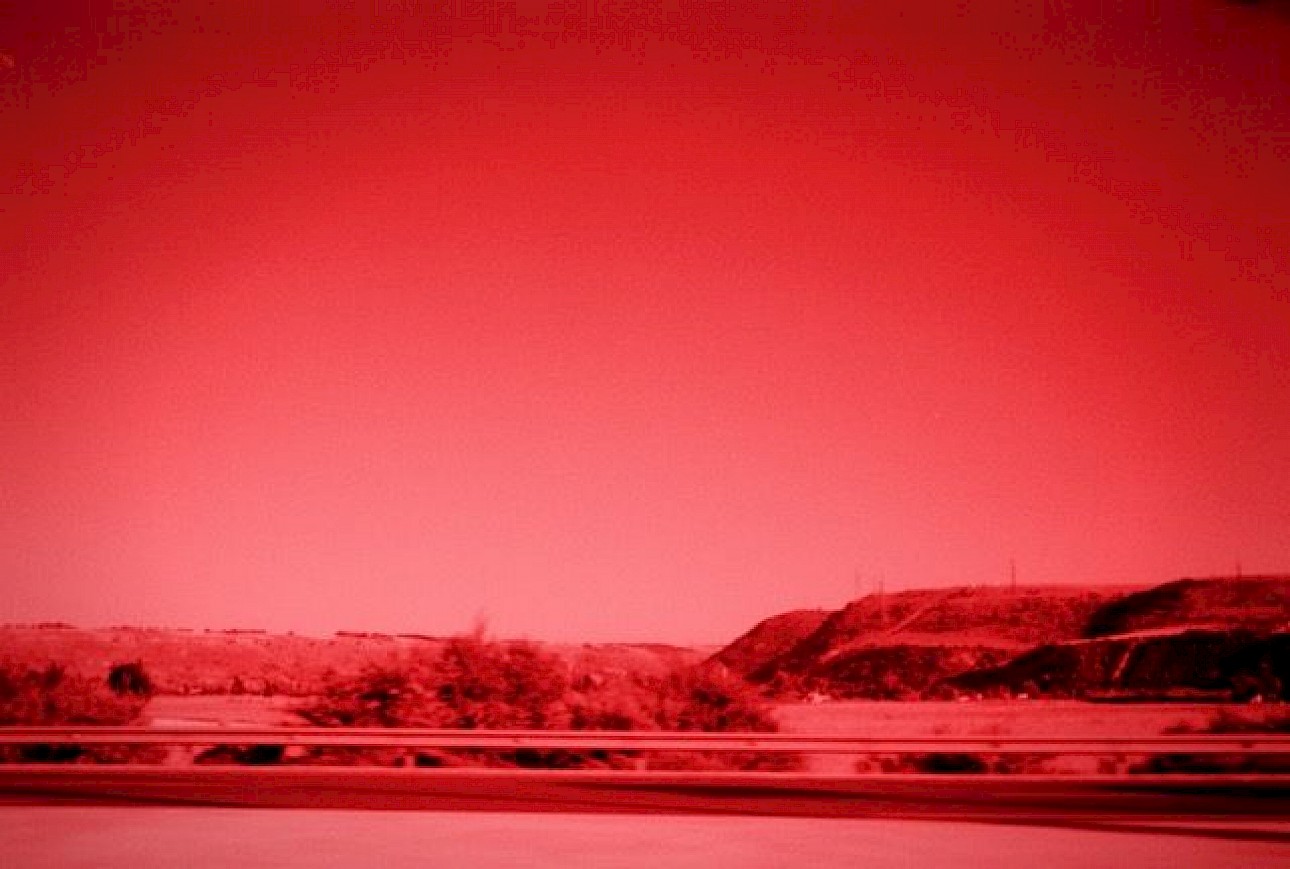 ALKANTARA - A view of a spot somewhere in the countryside, captured from a moving vehicle. We can see a bit of the road and the plains. The photograph is in shades of red and black. - ©Amel Malfait Lakhtara