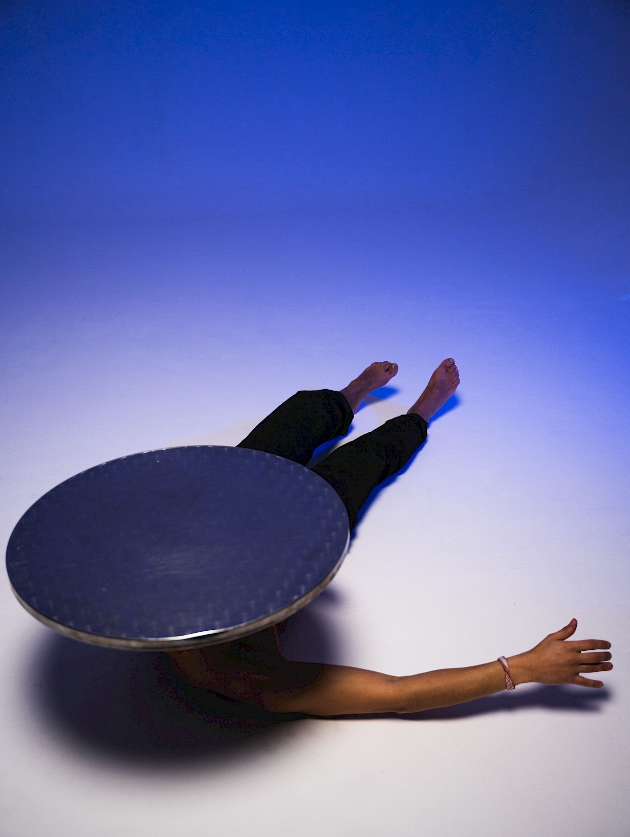 ALKANTARA - Overhead view of a person lying on their back on a white floor under blue lighting, wearing dark trousers and barefoot. Their upper body and head are hidden underneath a large round metal tabletop. - ©Njaheut Valler