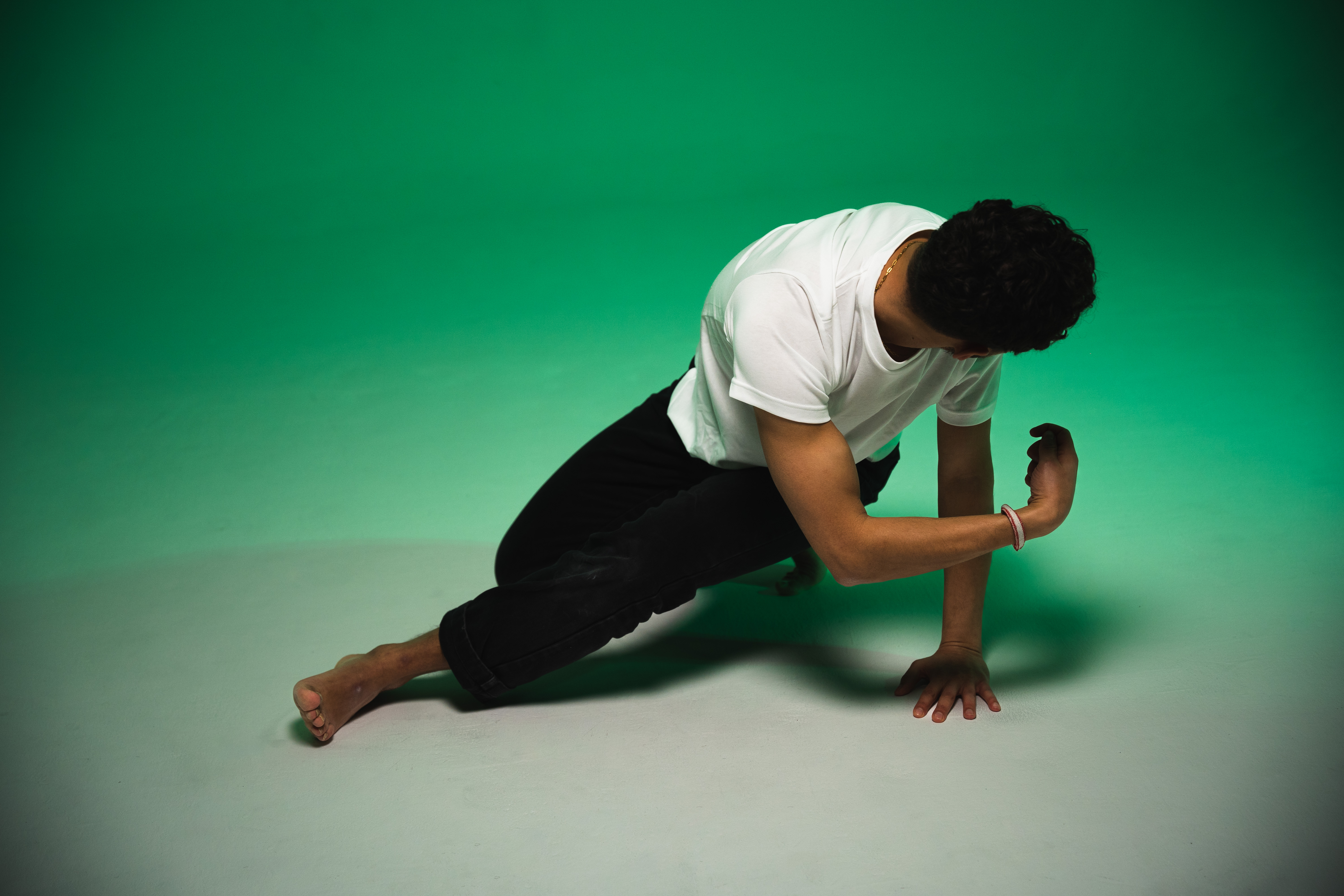A barefoot man in a white t-shirt and black trousers crouching in a dance pose against a green studio backdrop. His body is leaning forward with one hand resting on the floor and his head turned away. - Njaheut Valler