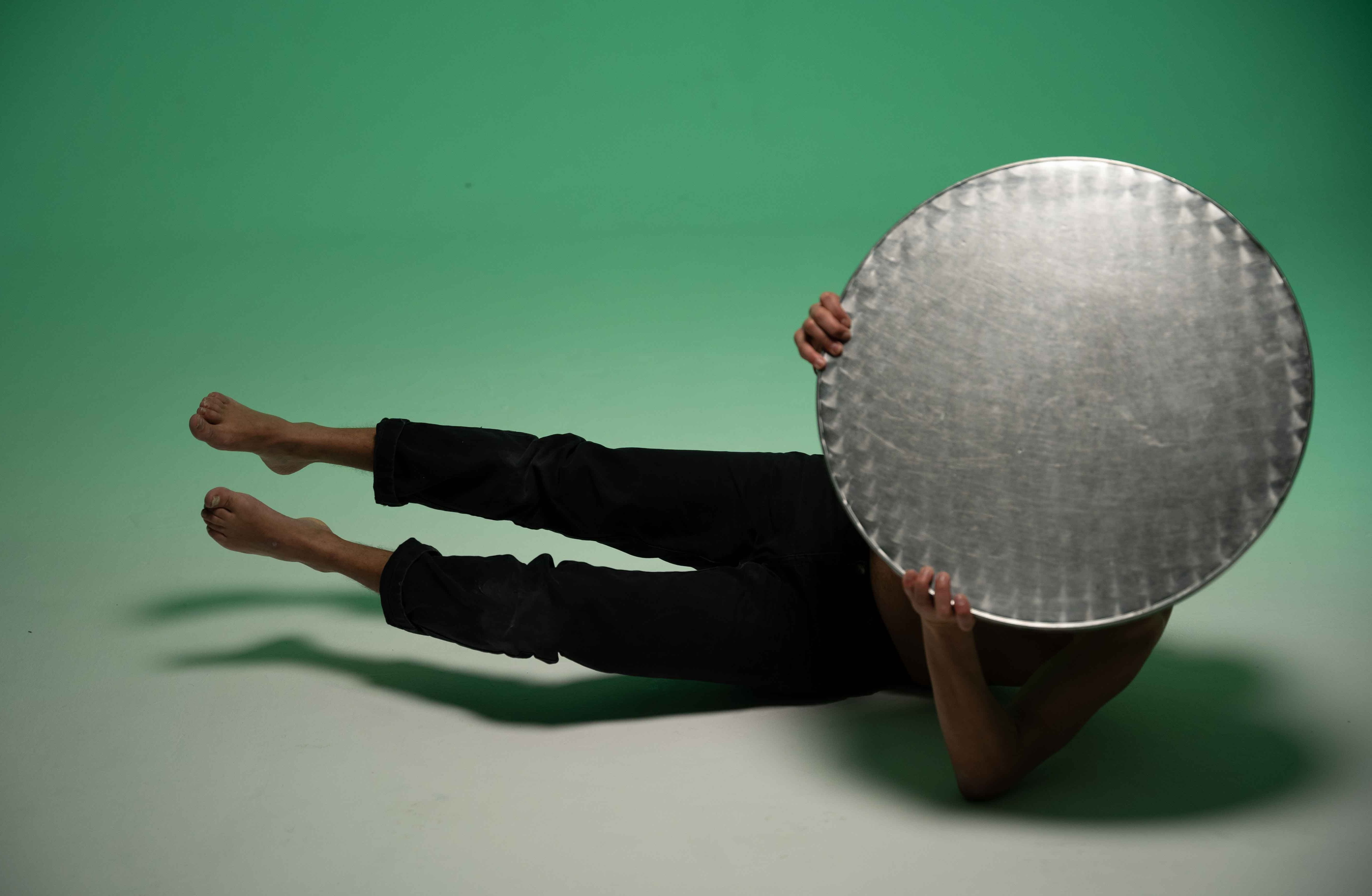 A person lying on their side on a light floor against a green backdrop, holding a large round metal disc that obscures their face and upper torso. Their bare legs extend out to the left. - Njaheut Valler