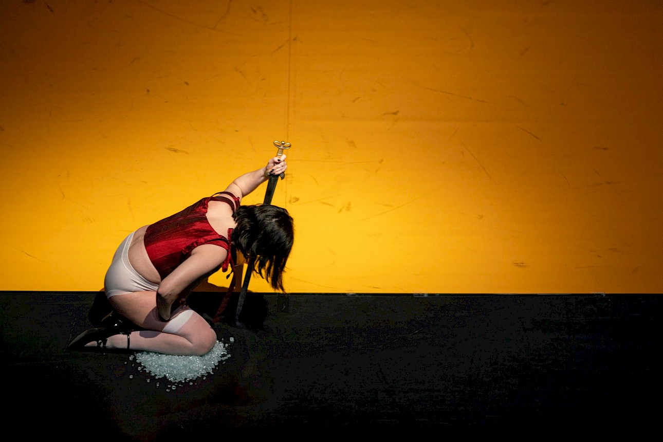 ALKANTARA - High-angle shot of a performer kneeling on a small pile of light-coloured stones or glass on a stage split between a black floor and a vibrant yellow backdrop. She wears a dark red corset, light underwear, white stockings, and high heels, holding a dagger pointed downward near her neck with her head bowed. - ©Mayra Azzi