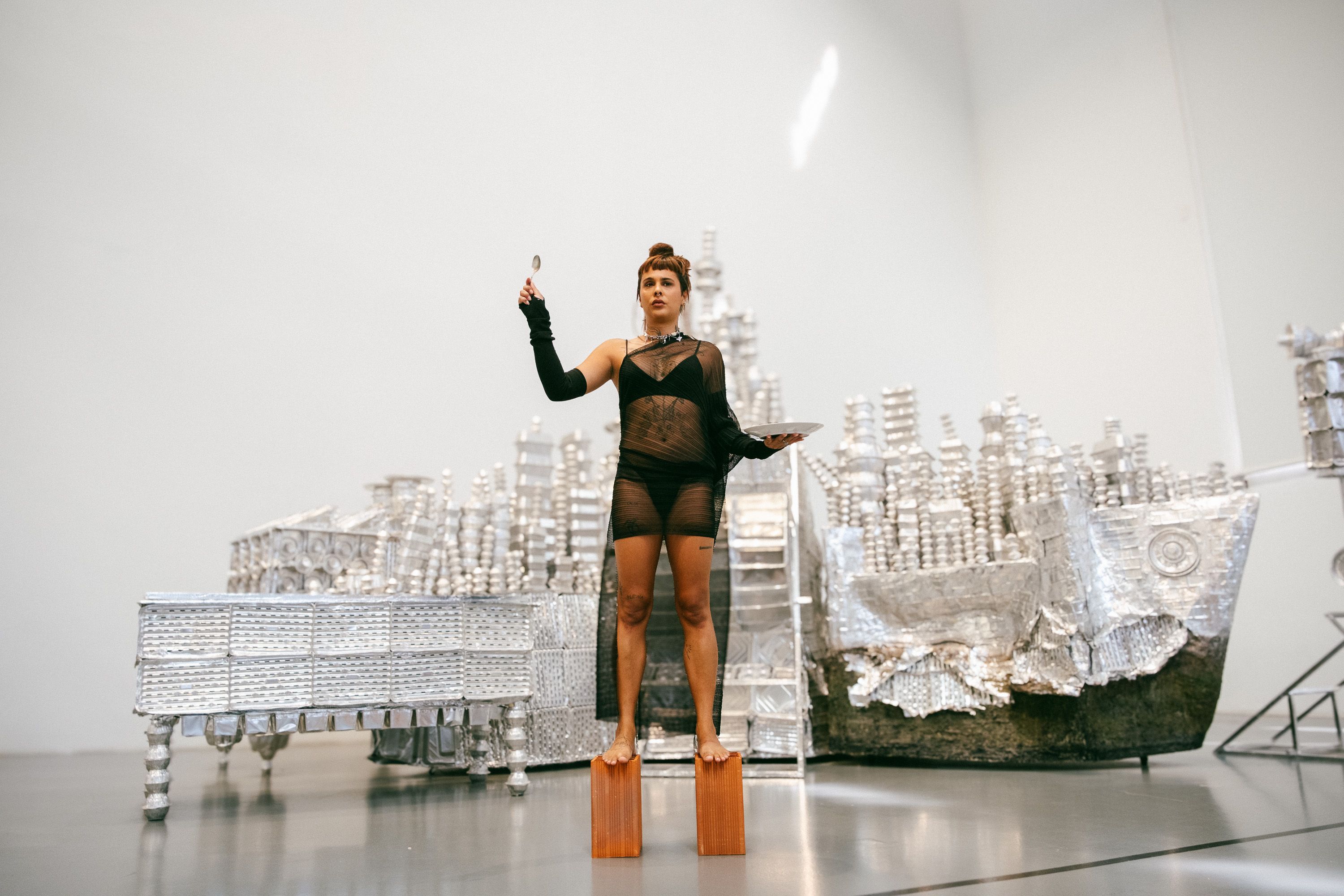 Vista geral de uma artista equilibrada em cima de dois blocos de madeira num espaço amplo de galeria de arte. Ela veste um fato/vestido preto transparente, segura uma colher erguida e um prato. Atrás dela encontra-se uma grande instalação artística prateada feita de recipientes de alumínio que lembram edifícios e um barco. - Diana Mendes