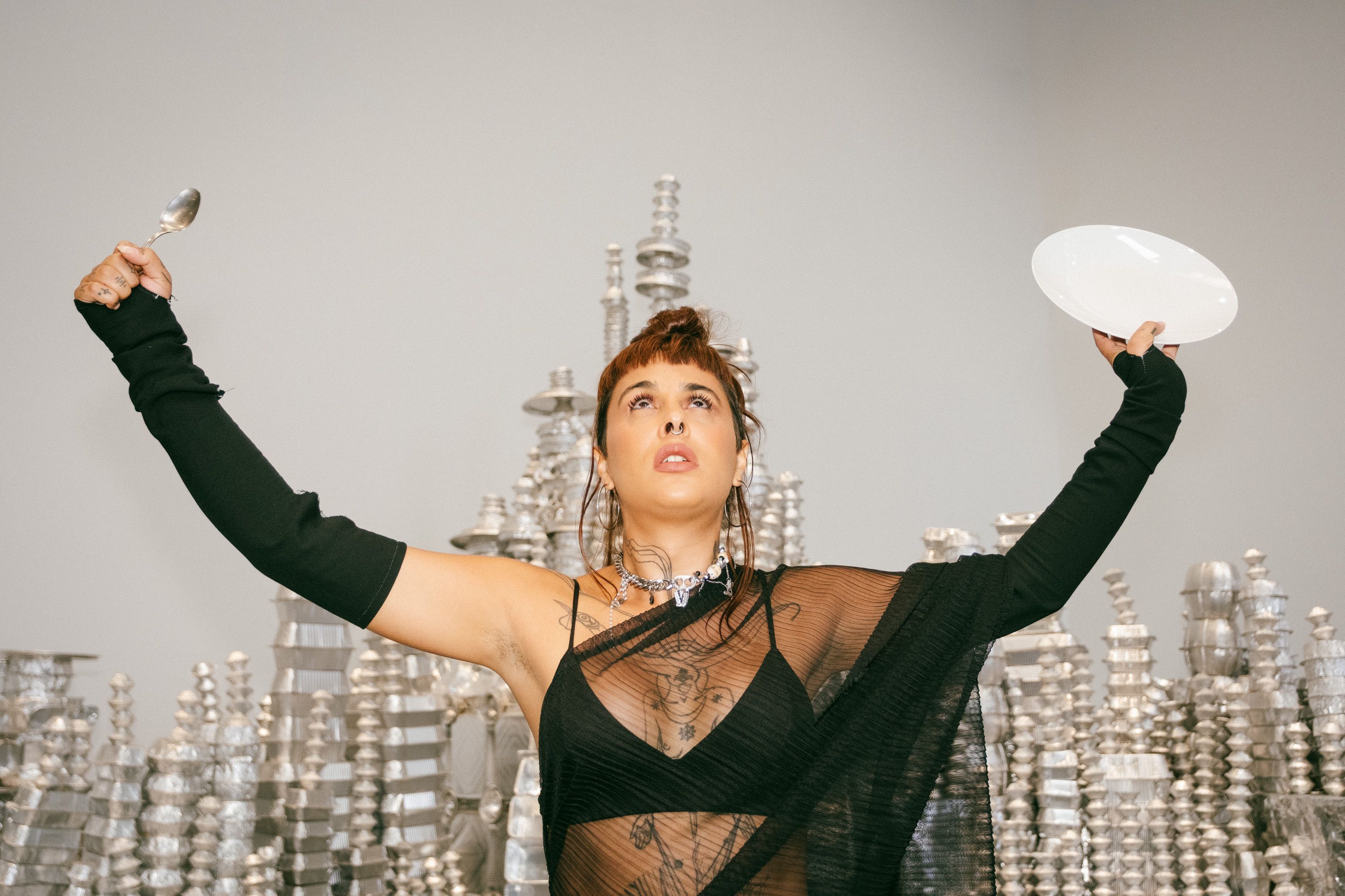 Plano aproximado de uma performer com cabelo curto castanho e franja, usando um vestido preto transparente e luvas longas. Ela olha para cima com os braços erguidos em "V", segurando uma colher de metal na mão direita e um prato branco na mão esquerda. Ao fundo, veem-se estruturas prateadas e cintilantes empilhadas. - Diana Mendes