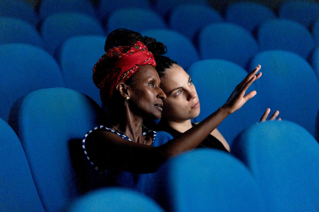 ALKANTARA - Medium shot of two people seated in blue cinema chairs. The woman in the foreground wears a patterned red headscarf and a blue top, pointing with an raised hand as she speaks to the person next to her, who listens intently. - ©Pietro Bertora
