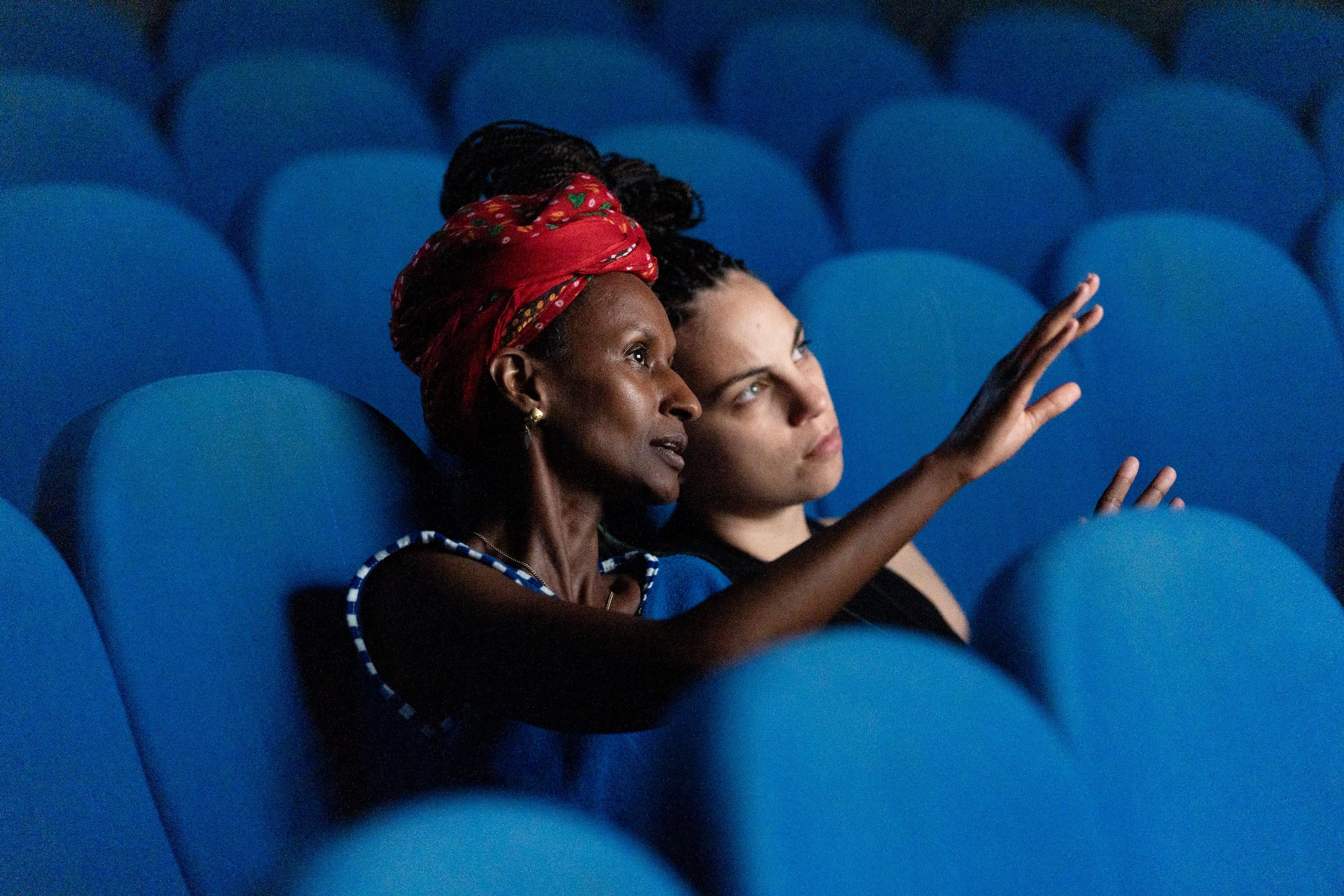 Medium shot of two people seated in blue cinema chairs. The woman in the foreground wears a patterned red headscarf and a blue top, pointing with an raised hand as she speaks to the person next to her, who listens intently. - Pietro Bertora