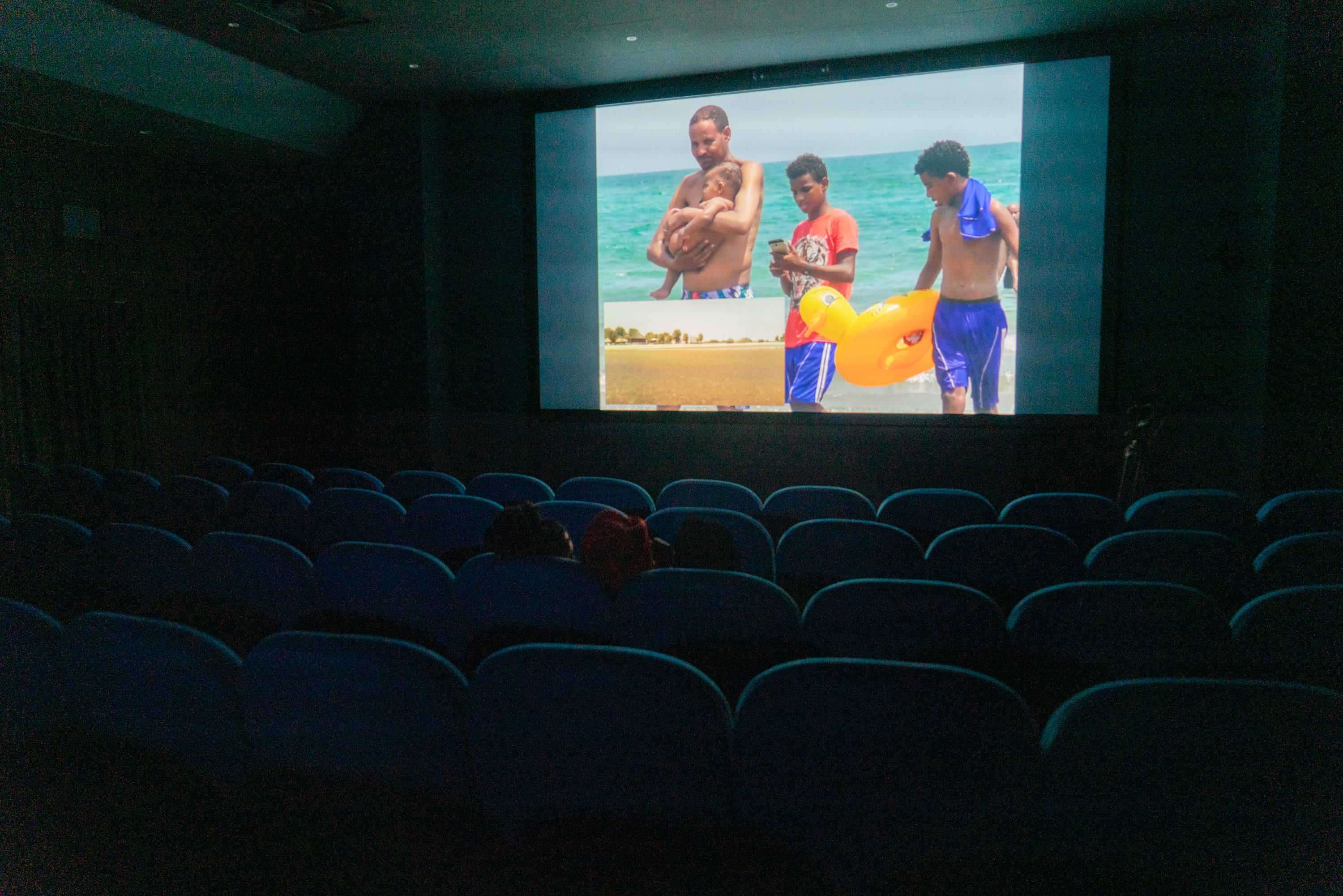 Wide shot of a dark cinema auditorium with rows of mostly empty blue seats. A colour photograph of several people on a beach by the sea is projected onto the screen at the front. - Pietro Bertora