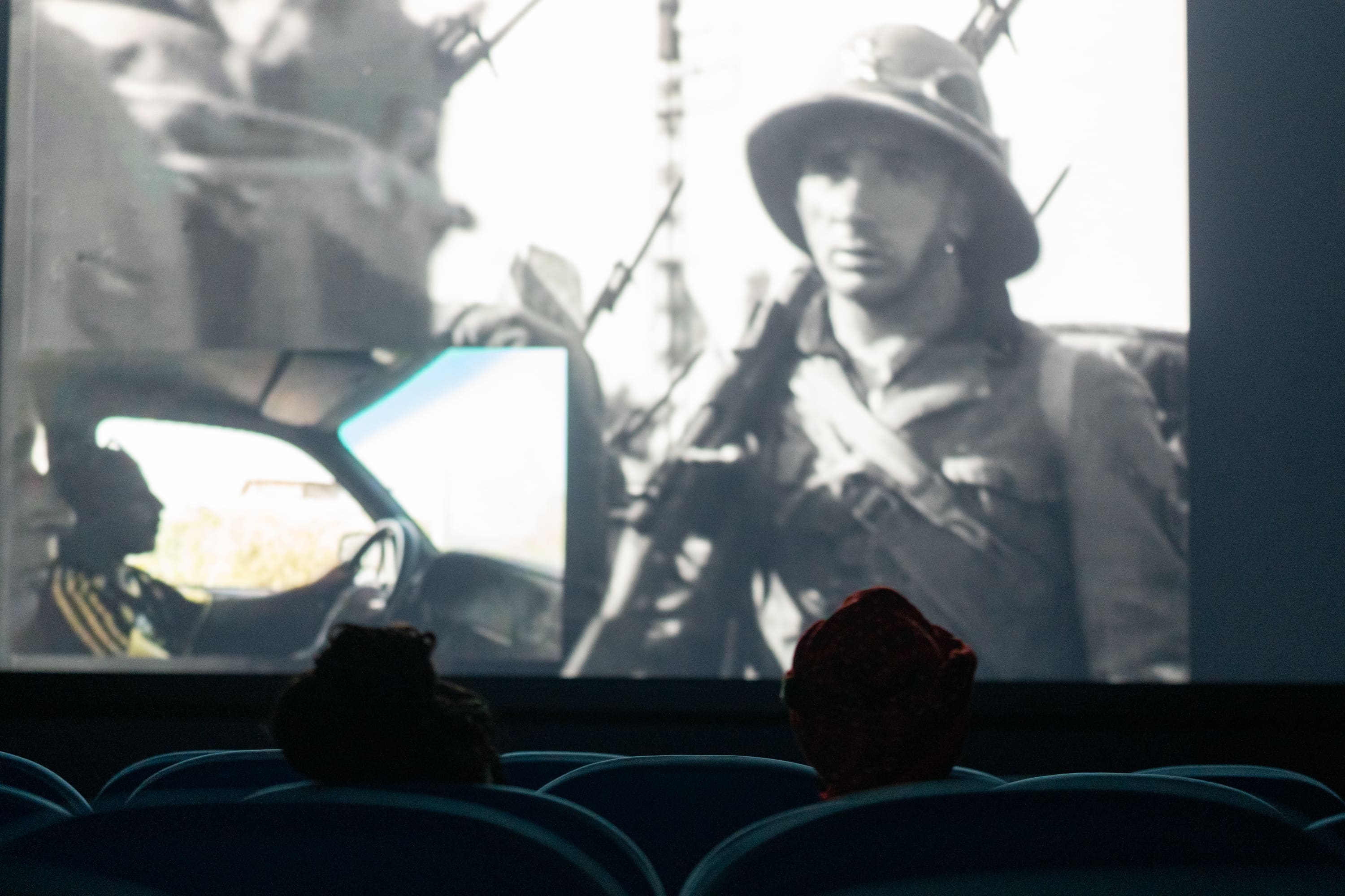 View from the back of a dark auditorium showing the silhouettes of two audience members in cinema seats, watching a film projection. The screen displays a historical black-and-white image of a helmeted soldier with a smaller picture-in-picture insert of someone driving. - Pietro Bertora