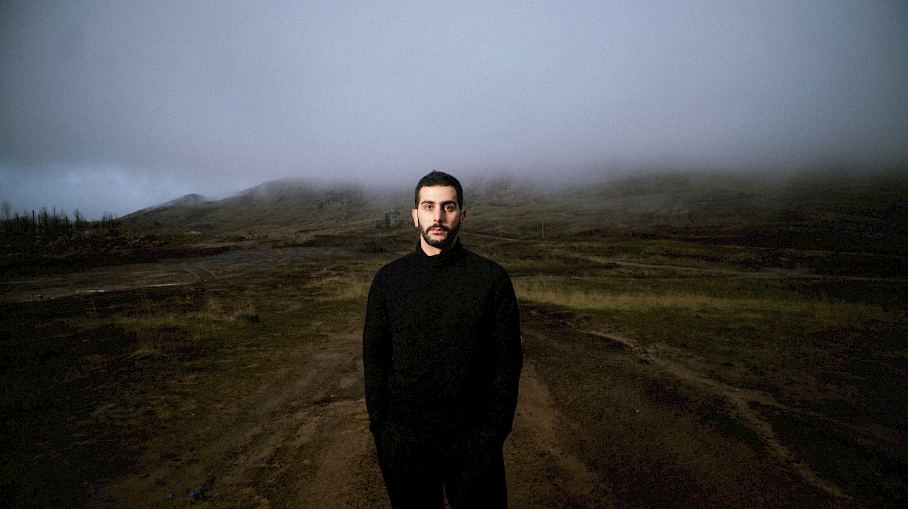 ALKANTARA - Retrato de Ali Chahrour. Plano médio de um homem de barba curta e cabelo escuro, vestido com uma camisola de gola alta e calças pretas, posando de frente num caminho de terra. Ao fundo, estende-se uma paisagem rural e montanhosa sob um céu nublado e com névoa baixa. - ©Lea Skayem