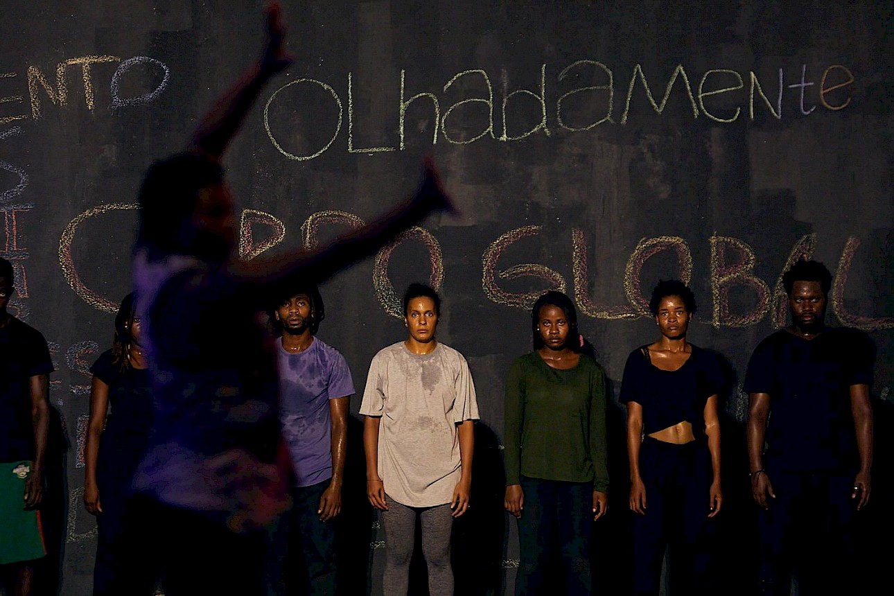 ALKANTARA - Em primeiro plano, uma figura desfocada gesticula com os braços erguidos em direção a um grupo de seis performers alinhados no centro de um espaço de ensaio. Os performers estão de pé com olhar concentrado, vestindo roupa casual e desportiva, em frente a uma parede escura coberta de frases e palavras escritas a giz. - ©Mariano Silva