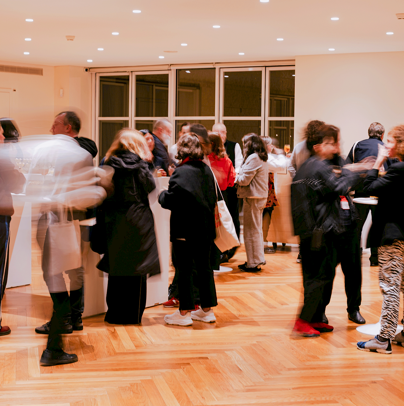 ALKANTARA - Pessoas convivem e conversam num evento social dentro de uma sala iluminada com chão de taco de madeira e teto com focos embutidos. Algumas pessoas surgem ligeiramente desfocadas devido ao movimento da fotografia. Ao fundo, veem-se janelas de vidro de grande dimensão. - ©Beatriz Pequeno