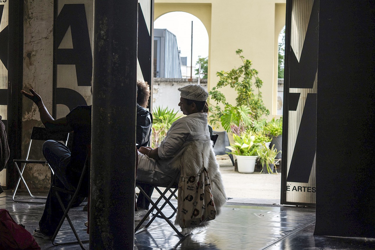 ALKANTARA - People sitting on folding chairs inside an indoor space with pillars and open glass doors. In the foreground, a person gestures with an raised hand while speaking; in the center, another person wearing a light beret and a fluffy coat draped over their chair looks outside. In the background, an outdoor courtyard features potted plants and architectural arches. - ©Rui Palma