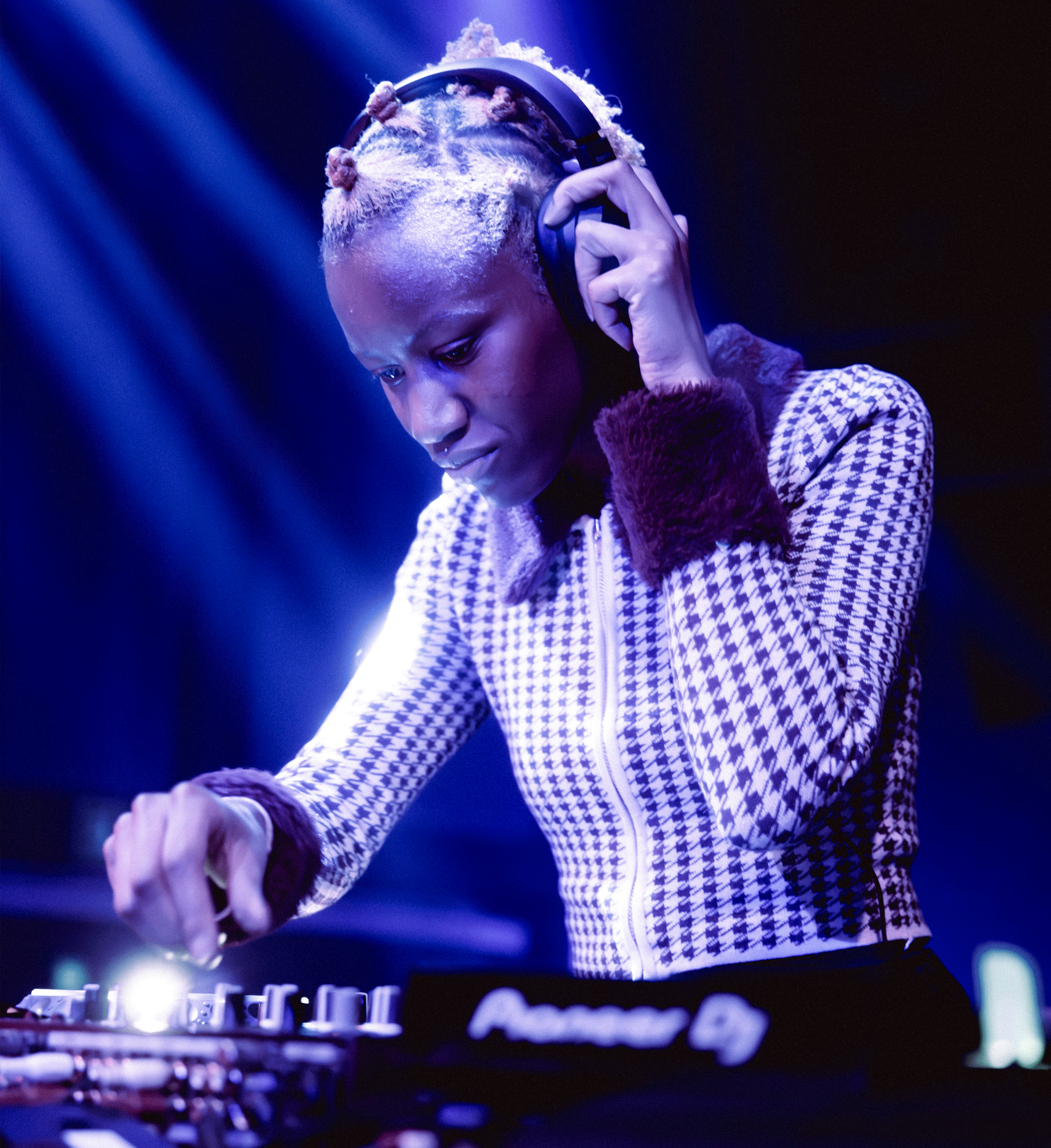 Medium shot of a Black DJ with bleached hair styled in small topknots, focused on mixing music on a DJ console under blue lighting. They hold headphones to one ear and wear a houndstooth patterned jacket with velvet trim on the cuffs and collar. - tomicornio