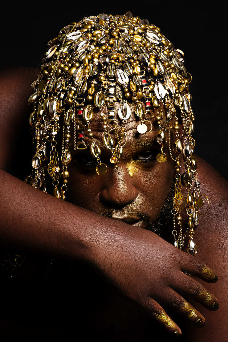 © Feeh Garcia DIDI's portrait. Close-up portrait of a Black person with gold makeup on their face, wearing an elaborate headpiece made of cowrie shells and gold pendants. - Feeh Garcia