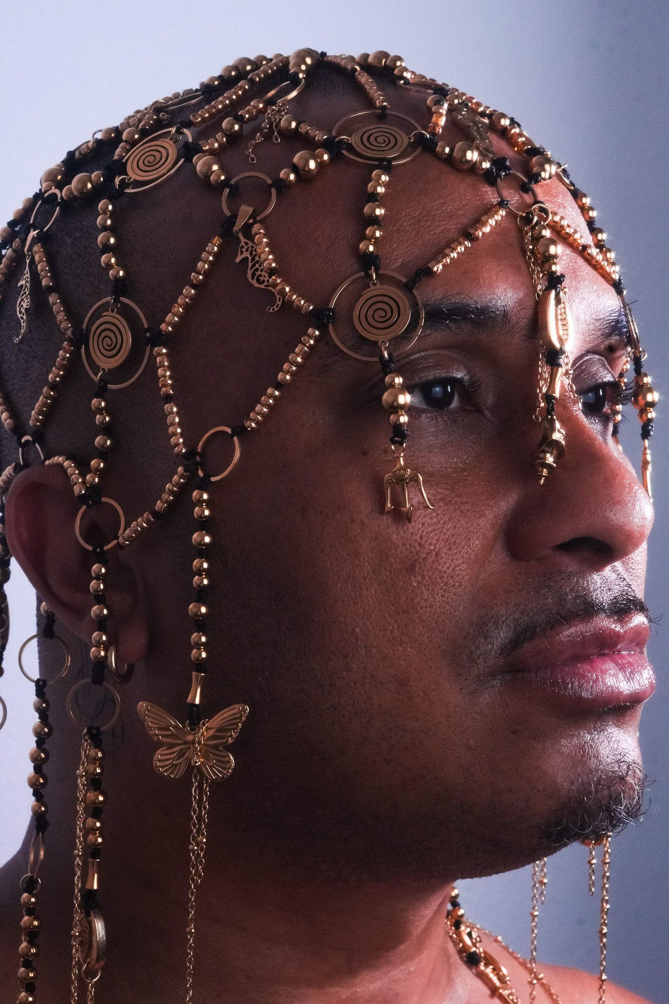 © Joni Ricos Profile portrait of a Black person wearing an intricate gold headpiece with beads, chains, and charms, including a butterfly. - Joni Ricos