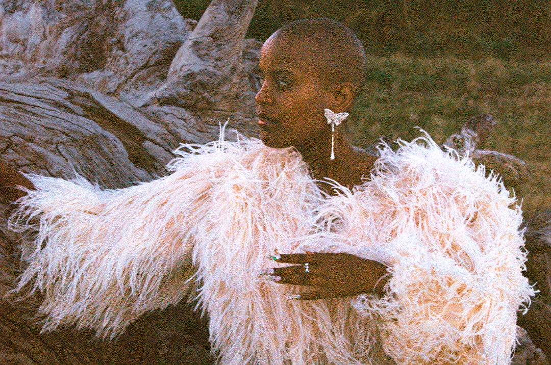 © Raye Kreidel Profile portrait of a Black person with a shaved head wearing a voluminous beige feather jacket and a silver butterfly earring. Their hand is resting against an old tree trunk outdoors, showcasing long decorated fingernails. - Raye Kreidel