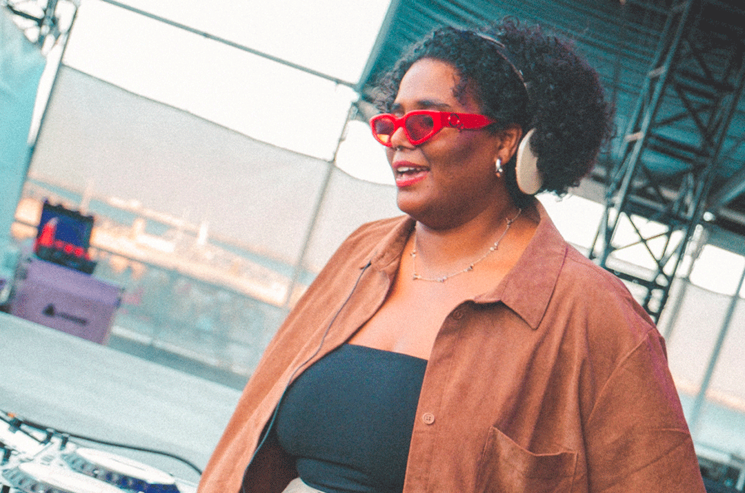 A smiling DJ wearing red sunglasses and white headphones around her neck, standing behind a DJ booth. She wears an open brown shirt over a black top on an outdoor stage beneath a metallic structure.