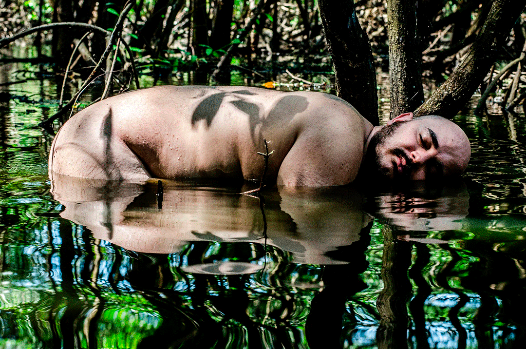 © Luiz Alves A bare-chested person with a shaved head floating in the calm water of a dense mangrove or swamp, surrounded by trees and vegetation reflected on the water's surface. - Luiz Alves