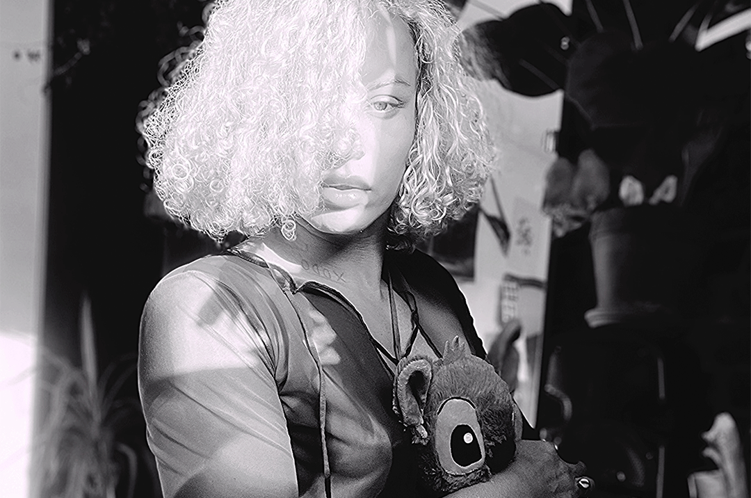 Black and white photograph of a person with short, voluminous curly hair looking directly at the camera while holding a small soft toy against their chest.