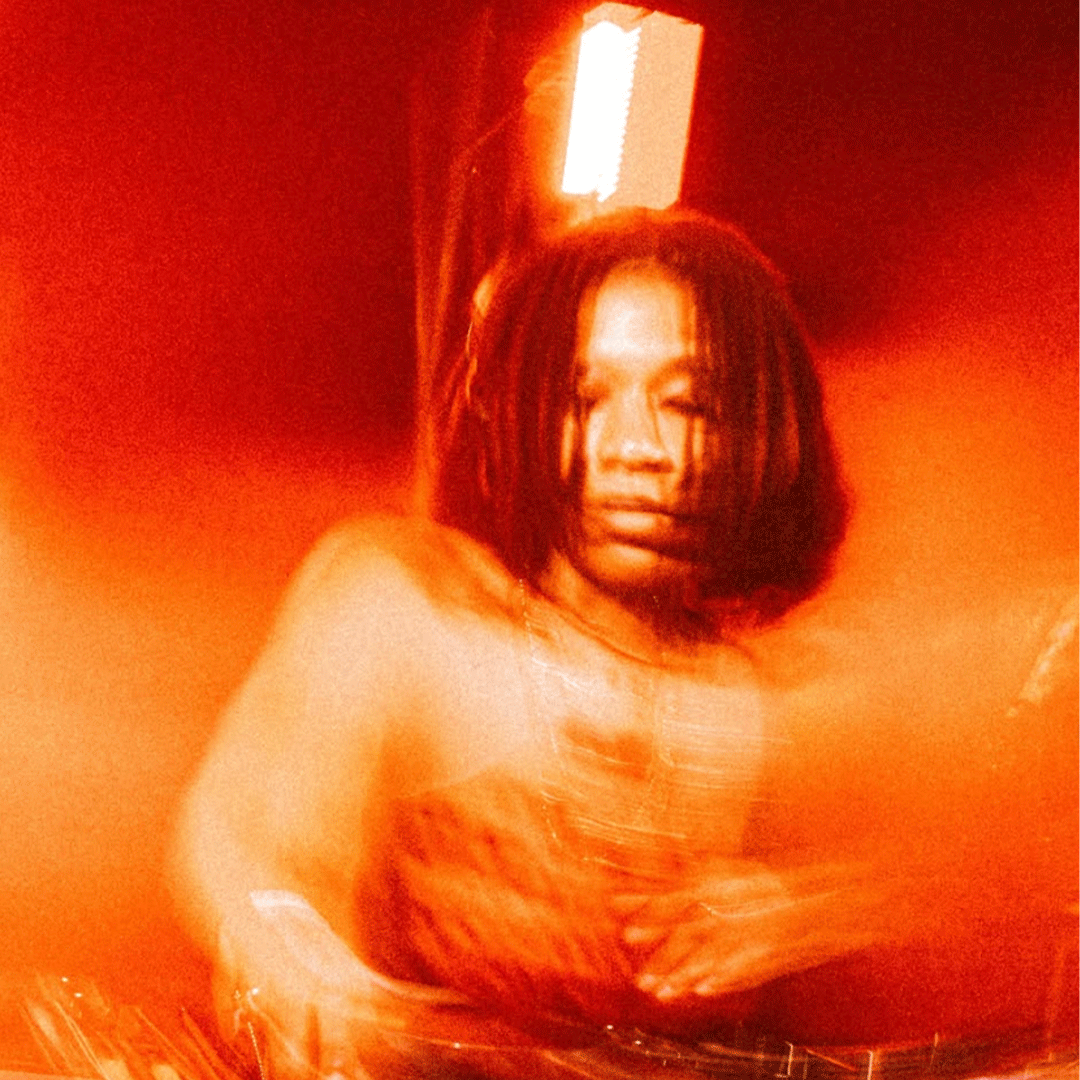 Motion-blurred portrait in vibrant red and orange tones, showing a person with short dreadlocks performing as a DJ behind a mixing console with warm lighting in the background.