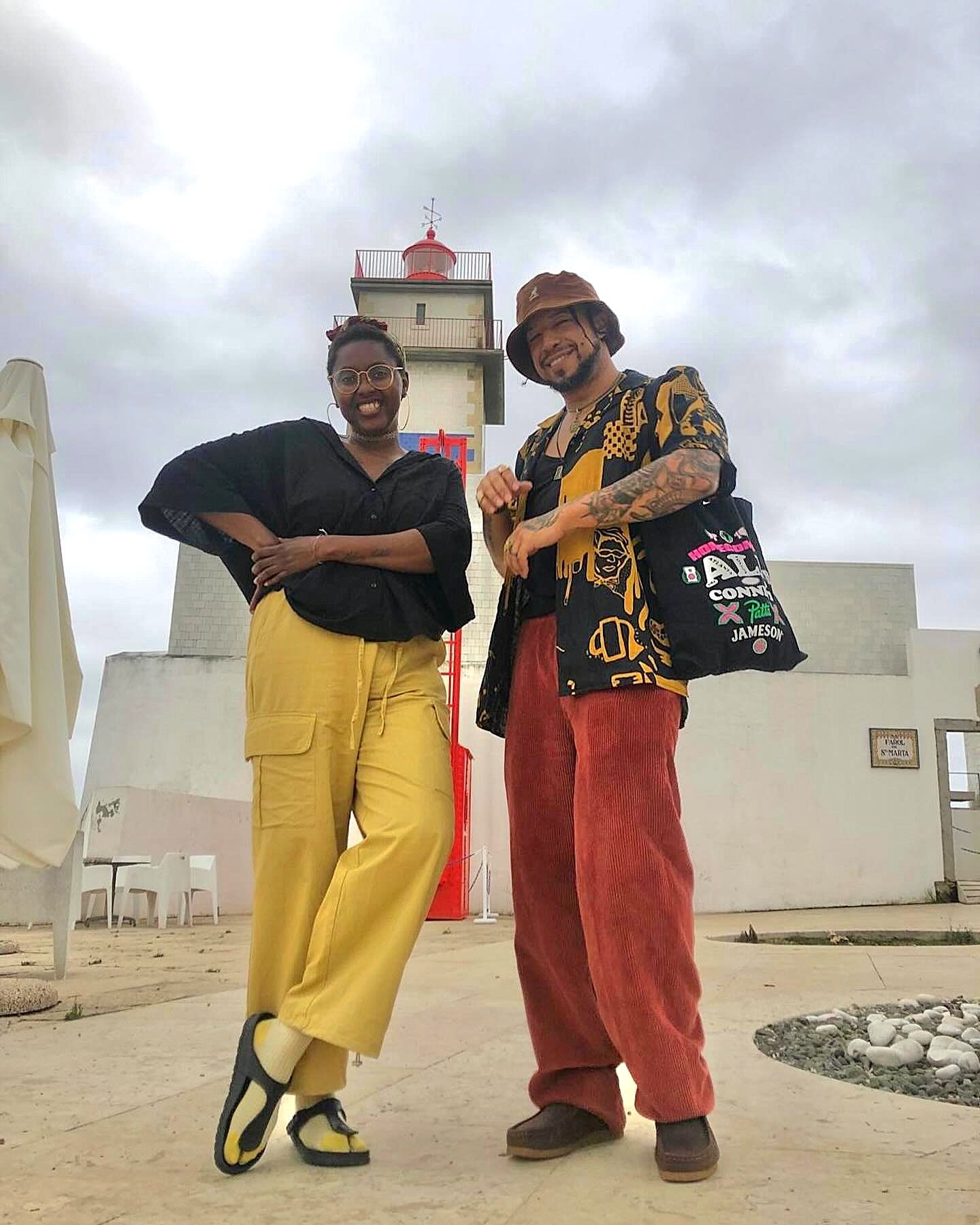 Two people pose smiling outdoors in front of a lighthouse under an overcast sky. The person on the left wears a black top, yellow cargo trousers, and round glasses, posing with one hand on their hip. The person on the right wears a brown bucket hat, a patterned black and yellow short-sleeved shirt over a black top, wide-leg terracotta corduroy trousers, and carries a tote bag over their shoulder.