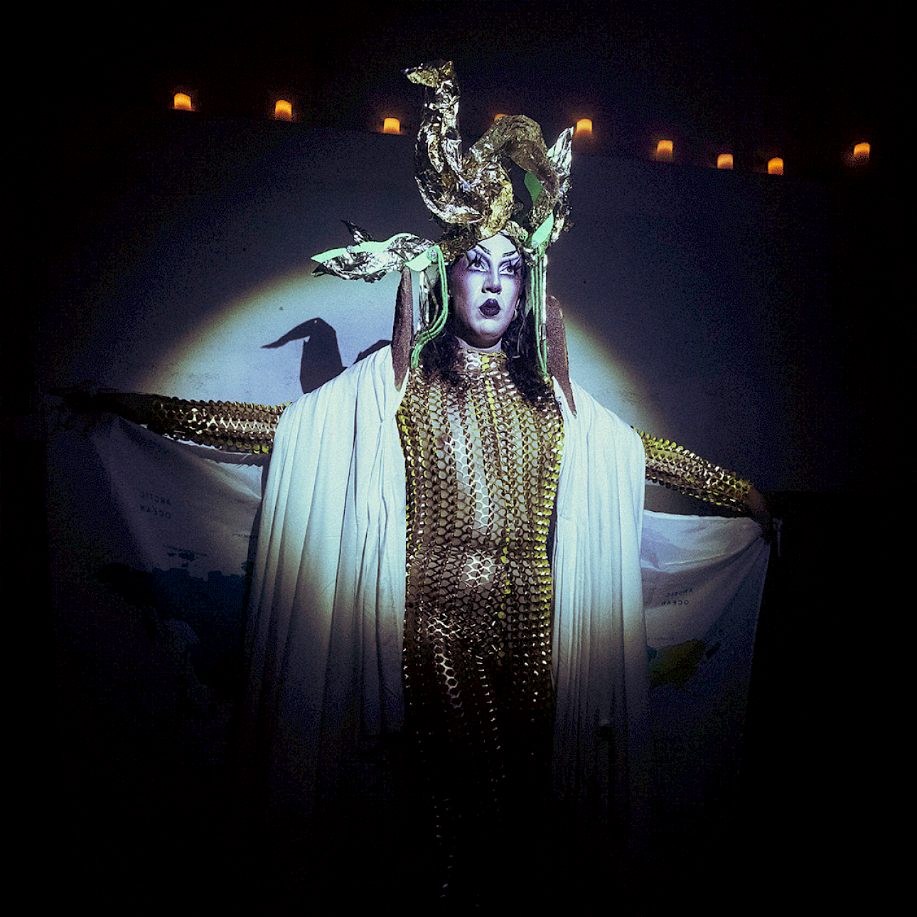 ALKANTARA - Performing artist wearing a fitted gold sequin mesh bodysuit and a draped white cape, posing with arms outstretched on a dark stage under a spotlight. They wear an elaborate gold headpiece and dramatic makeup, holding what appears to be a world map printed on fabric. - ©tomi cornio