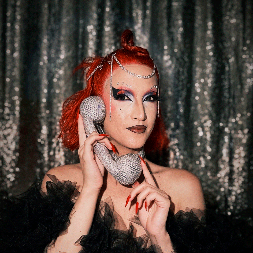 Medium portrait of a performer with bright red hair styled in an updo and elaborate drag makeup, holding a glittering silver vintage telephone handset to their ear. They wear a black ruffled tulle outfit, long red nails, and silver head chains, posed in front of a shimmering silver sequin backdrop.