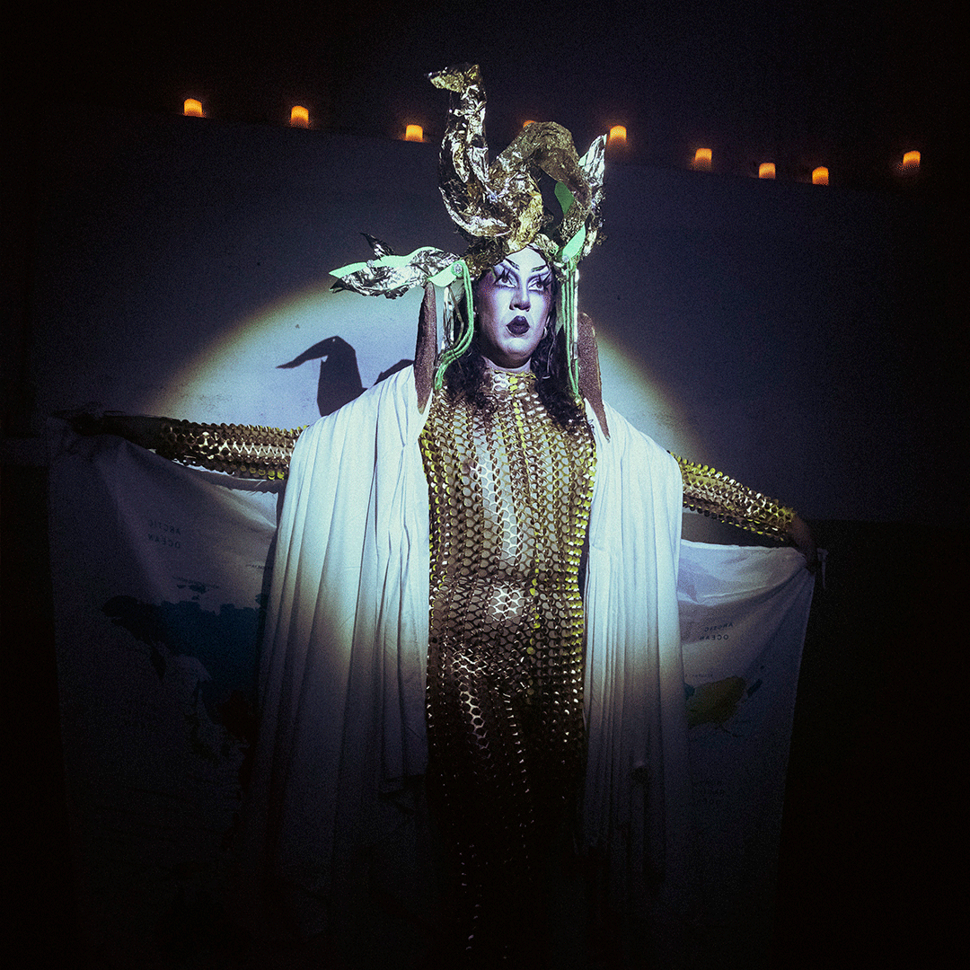 Performing artist wearing a fitted gold sequin mesh bodysuit and a draped white cape, posing with arms outstretched on a dark stage under a spotlight. They wear an elaborate gold headpiece and dramatic makeup, holding what appears to be a world map printed on fabric. - tomi cornio